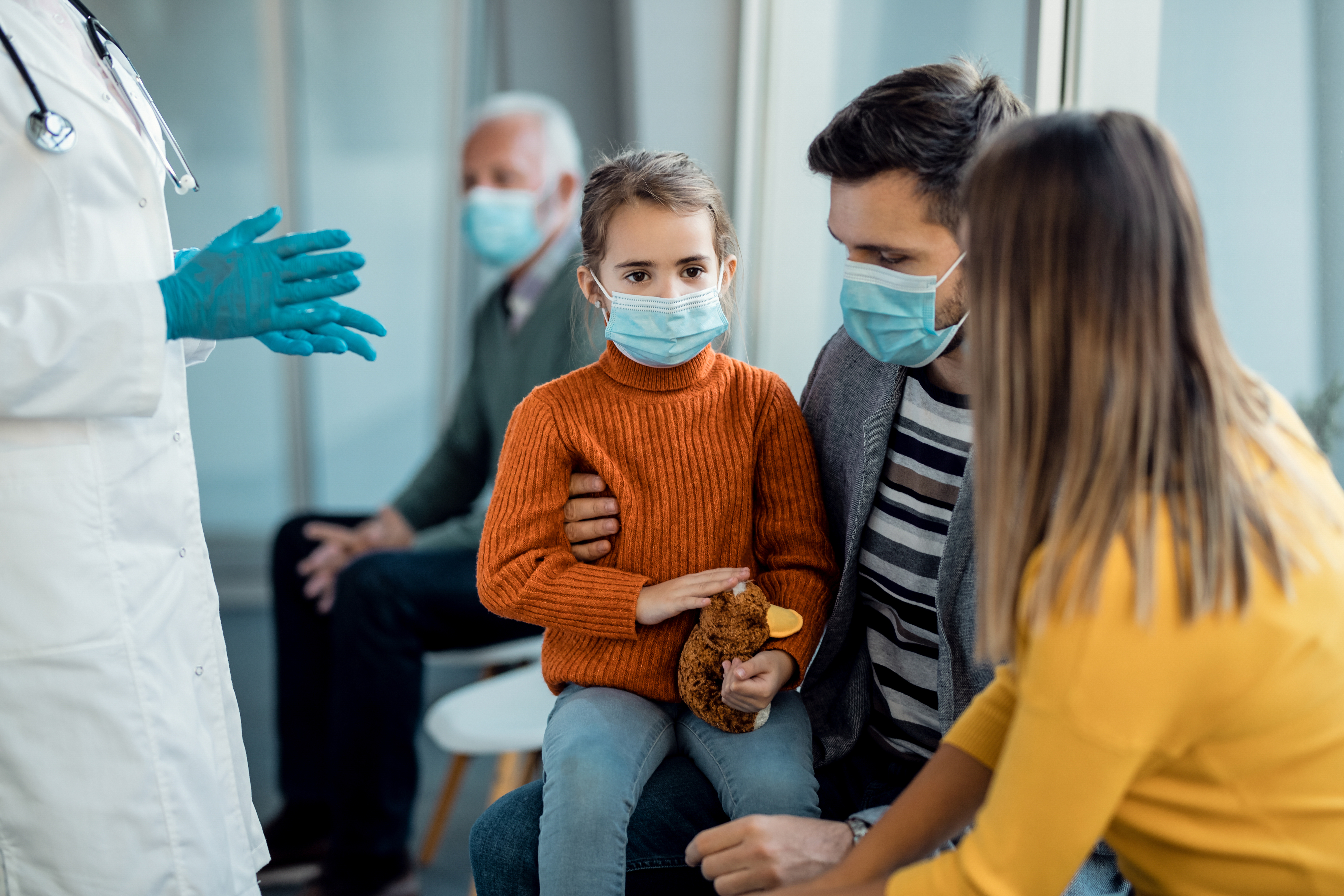 Small girl and her parents wearing protective face masks while sitting in a waiting room at medical clinic.